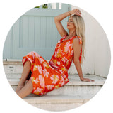 Model sitting in front of a gate wearing an orange floral dress. Links to the dresses collection.