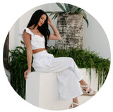 Model sitting on a wall wearing a white ruffle two piece set. Links to the sets collection.